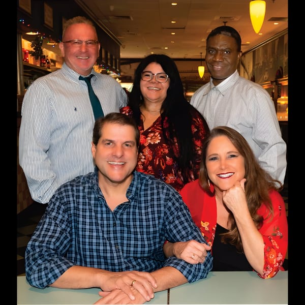 Five smiling people, three men and two women, posing together in a restaurant setting.