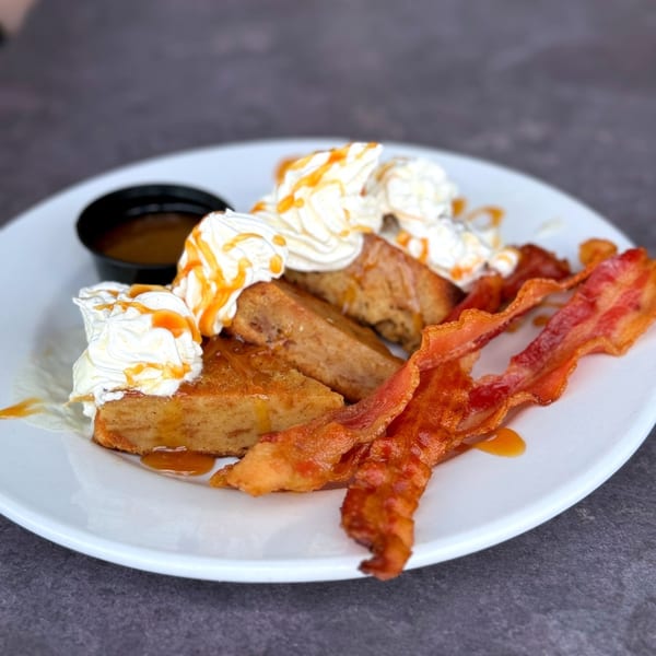 French toast with whipped cream, caramel drizzle, and crispy bacon on a white plate.