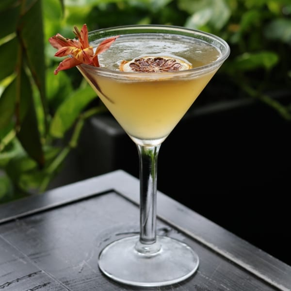 A martini glass with a yellow cocktail, garnished with a dried citrus slice and an orange flower.