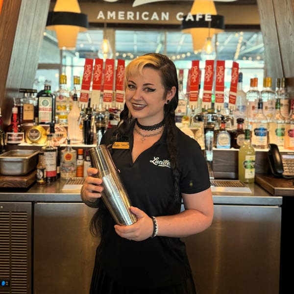A smiling Lucille's American Cafe bartender holds a cocktail shaker behind a well-stocked bar.