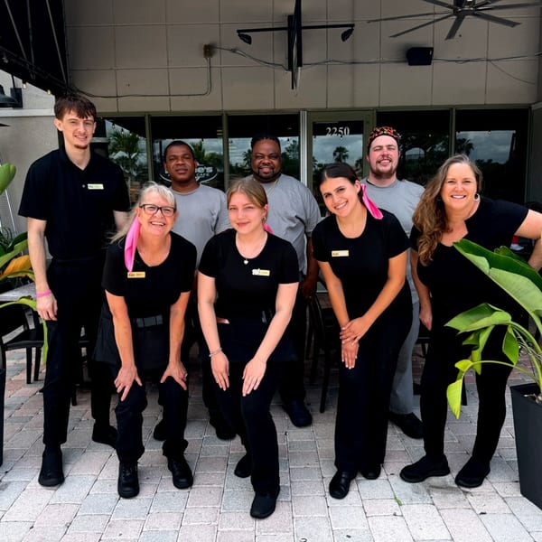 Eight smiling Lucille's American Cafe staff members, some wearing pink ribbons, pose outside the restaurant.