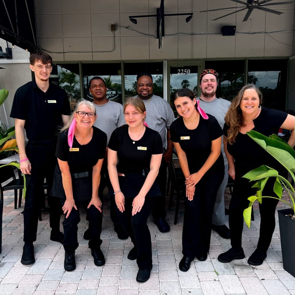 Eight smiling Lucille's American Cafe staff members, some wearing pink ribbons, pose outside the restaurant.