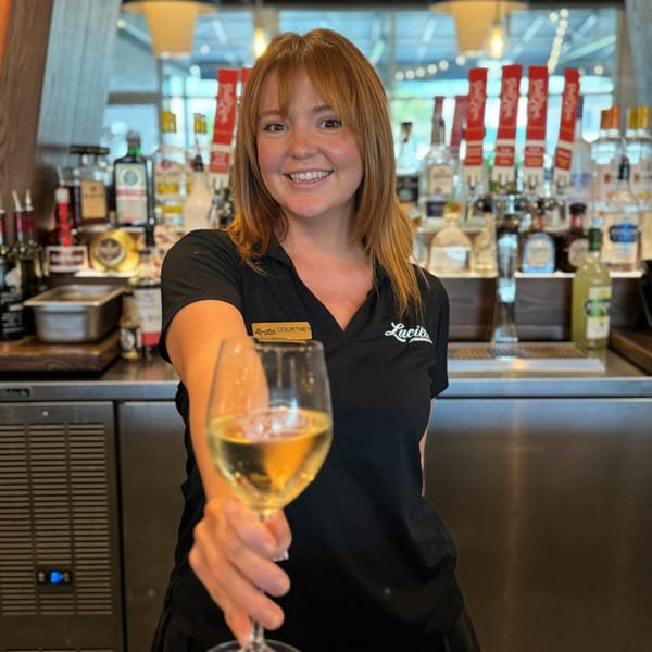 A smiling Lucille's bartender in a black polo shirt holds out a glass of white wine.