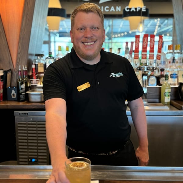 A smiling Lucille's bartender in a black polo shirt serves a cocktail with a citrus slice.