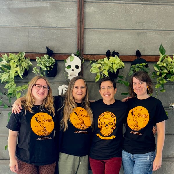 Four smiling women in Lucille's American Cafe t-shirts stand in front of a plant-decorated wall.